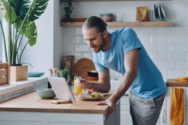 Yakışıklı adam yemek yiyor ve mutfak tezgahında dikilirken dijital tablete bakıyor.