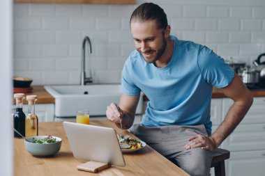 Evde mutfak tezgahında otururken yemek yiyen yakışıklı genç adam.