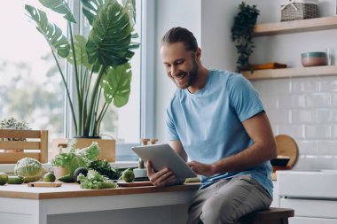 Mutfakta yemek hazırlarken elinde dijital tablet tutan neşeli genç adam.