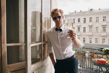 Yakışıklı genç adam balkon kapısında eğilmişken şampanya ile kadeh kaldırıyor.