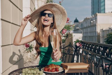 Şampanyayla kadeh kaldıran ve balkonda dinlenirken gülümseyen zarif şapkalı güzel bir genç kadın.