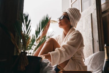 Bornozlu güzel bir kadın balkonda dinlenirken şampanya içiyor.