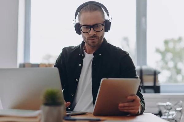 坐在办公室的工作位置时 一个拿着耳机用技术忙碌的年轻人 — 图库照片