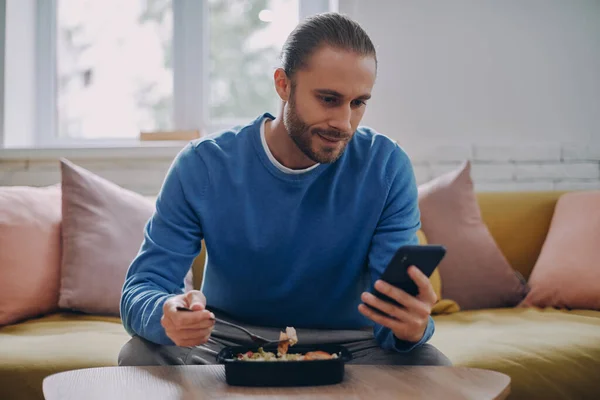 Yakışıklı Genç Adam Yemek Yiyor Evde Kanepede Otururken Akıllı Telefon — Stok fotoğraf