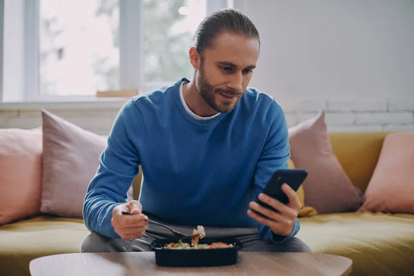 Bonito Jovem Almoçando Usando Telefone Inteligente Enquanto Sentado Sofá Casa — Fotografia de Stock