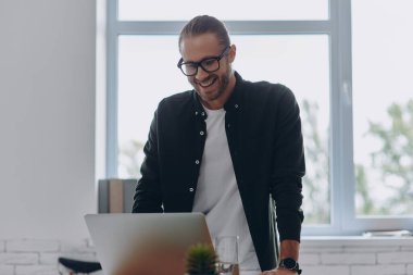 Ofisteki iş yerinin yanında dizüstü bilgisayar kullanan mutlu genç adam.