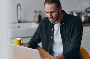 Evde mutfak masasında çalışırken dizüstü bilgisayar kullanan yakışıklı bir genç.