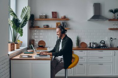 Kulaklıklı neşeli genç adam evde mutfak tezgahında otururken dizüstü bilgisayarla çalışıyor.