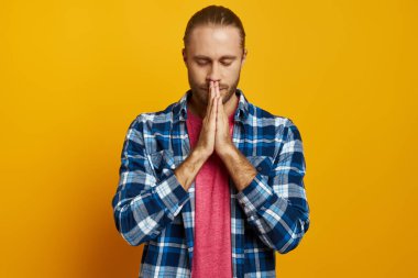 Sarı arka planda dikilen genç adam parmaklarını çapraz yapıp gözlerini kapatıyor.