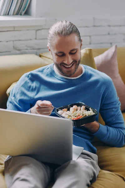 Relaxed young man eating lunch and using laptop while sitting on the couch at home
