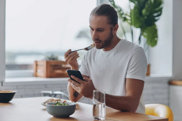 Genç Adam Yemeğin Tadını Çıkarıyor Mutfakta Otururken Akıllı Telefon Kullanıyor — Stok fotoğraf