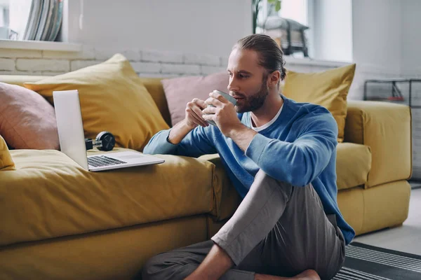 Guapo Joven Disfrutando Bebida Caliente Mirando Computadora Portátil Mientras Está — Foto de Stock