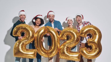 Altın renkli numaralar taşıyan ve gri arka planda dururken gülümseyen bir grup genç Noel şapkalı güzel insan.