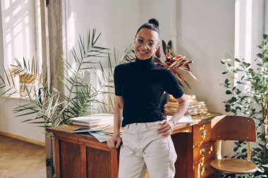 Güzel bir kadın iş yerinin yanında dururken kameraya bakıyor ve gülümsüyor.