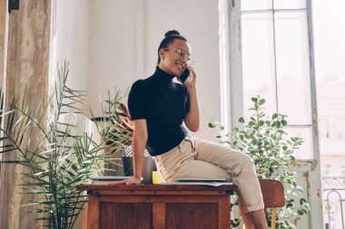 Evdeki ahşap masada otururken cep telefonuyla konuşan ve gülümseyen güzel bir genç kadın.