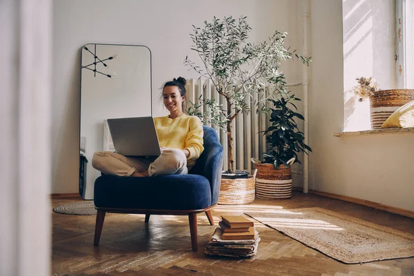 Gyönyörű Fiatal Használja Laptop Mosolyog Miközben Egy Kényelmes Székben Otthon — Stock Fotó