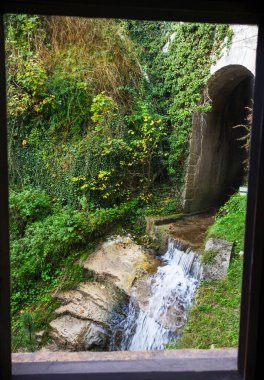 Bir pencere tarafından çerçevelenmiş açık hava manzarası. Taşlı bir kanalizasyondan akan tatlı su..