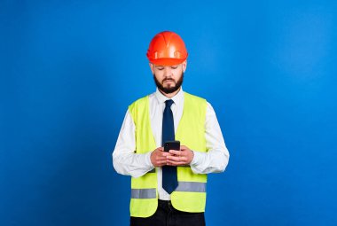 Koruyucu miğferli ve telefonlu yansıtıcı yelek giyen sakin genç bir inşaatçı. Mesajlaşma.