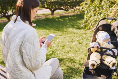 Modern, genç bir anne, bir sonbahar parkında küçük oğluyla bir bebek arabasında bankta oturuyor elinde akıllı bir telefonla, mesajlaşarak uzaktan çalışıyor. Annelik izni konsepti.