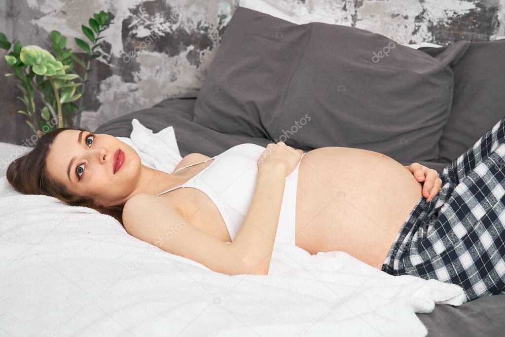 Pregnancy, rest, people. The concept of waiting for the birth of a child. Close-up of a frequent smiling pregnant woman lying in bed and touching her belly at home.