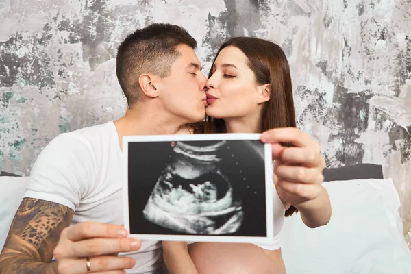 Genç ve sevecen bir çift, hamile bir kız, sabahları pijamalarıyla yatağa uzanıyor ve ultrason taramasında bir bebeğin resmini inceliyor..