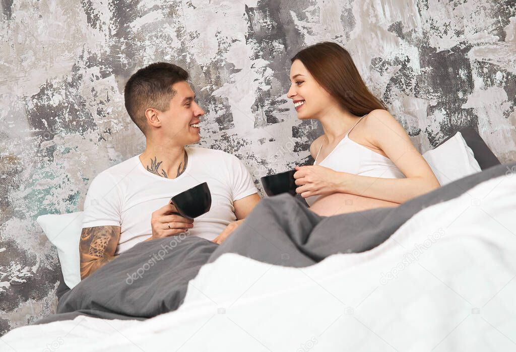 Happy tender young married couple in pajamas, drinking tea in bed and cheerfully discussing plans for the day. A young couple in anticipation of the birth of their first child. Family, love concept.