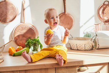 Neşeli, neşeli, yakışıklı küçük bir çocuk, 9 aylık, sarı pantolon ve çizgili tişört giymiş, anne yemek pişirirken mutfak masasında oturuyor. Çocuk elinde bir limon tutuyor..