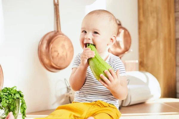 Sarı pantolonlu, çizgili tişörtlü sevimli, neşeli bir çocuk mutfak masasına oturmuş, elinde kabak tutuyor. Kabaklı beyaz bir çocuğun gülümseyen portresi..