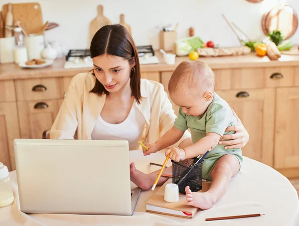 Cute caucasian young mother working on a laptop while sitting in the kitchen. Little boy plays with pencils, kitchen interior, free space. Working mom with baby, multitasking in the morning.