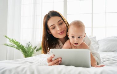 Anne ve küçük çocuk, birlikte yatak odasında oynuyorlar ve okuyorlar, evde tablet bilgisayarlar, sabahları güneş ışığıyla telefona bakıyorlar..