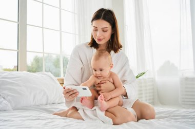 Beyaz bir yatakta küçük bir çocuk sabahları internet içeriğini taramak ve video izlemek için cep telefonu kullanıyor ya da ders çalışıyor. Yatakta bebeği olan genç bir anne..