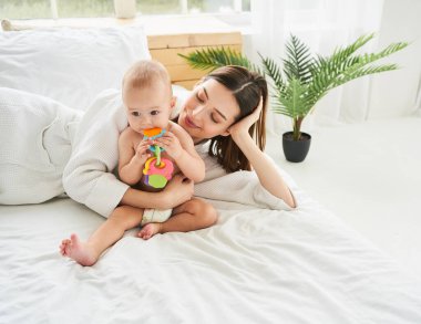 Altında bez olmayan sevimli bir erkek bebek annesinin yanındaki büyük beyaz bir yatakta güneşli bir sabahta oyuncaklarla oynuyor. Diş çıkarma, çocuk bakımı ve babalık kavramı..