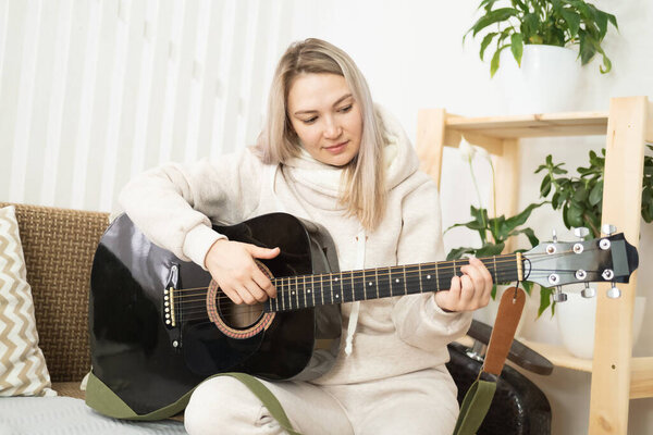 Beautiful middle aged blonde caucasian woman playing guitar while sitting on sofa at home. Home leisure, enjoyable pastime and relaxation