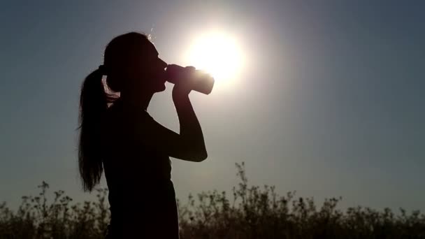 Jovem bebe água ao pôr do sol.Menina bebe água de uma garrafa (bancos ) — Vídeo de Stock