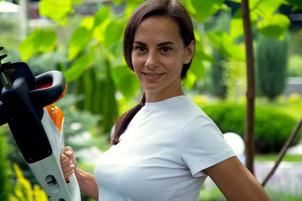Young beautiful happy woman holding garden tool looking at camera. Portrait of a beautiful gardener girl with a brush cutter. The concept of happiness, design, gardening, technology. 