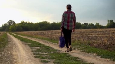 A young woman with a bag is walking along a country road. The girl returns home along a rural road with a bag in her hands. Camera movement behind the subject. The concept of loneliness, hopelessness. 
