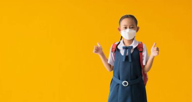 Asian student little girl wear medical face mask with show thumbs up for good, isolated on yellow background with Clipping paths for design work empty free space