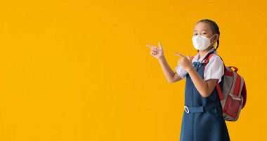 Asian student little girl wear medical face mask pointing fingers at copy space, isolated on yellow background with Clipping paths for design work empty free space
