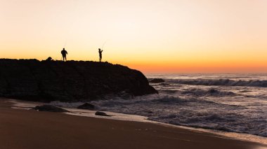Sahil kayalık kıyı şeridi sabah gökyüzüne bakan iki balıkçı silueti ile okyanus dalgaları manzarası .