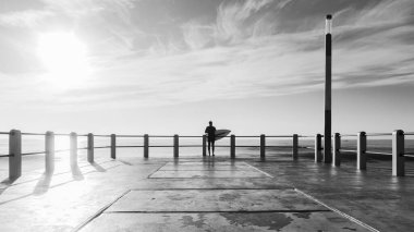 Sörfçü, sahildeki okyanus rıhtımının siluetinin ucunda okyanus suyuna atlamak üzere olan siyah beyaz bir fotoğrafın arkasında sörf yapıyordu..