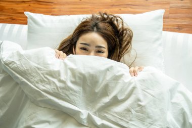 Yatakta beyaz battaniyenin altına saklanıp neşe dolu gözlerle kameraya bakan sevimli, genç bir kadın. Çekici kız yatak odasında eğleniyor..