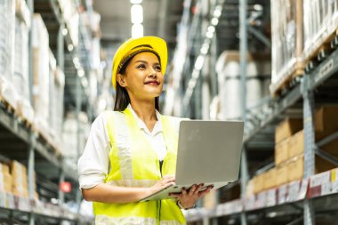 Gülümseyen Asyalı kadın depo çalışanı depodaki raflarda dizüstü bilgisayar, lojistik ve ticari ihracat kullanarak ürün yönetimini karton kutularda yapıyor.