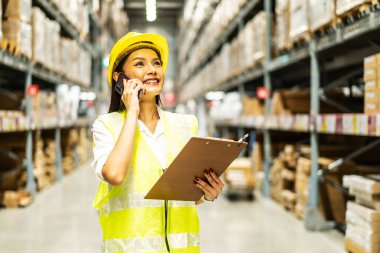 Miğferli ve üniformalı Asyalı kadın cep telefonuyla konuşuyor. Dağıtım deposu fabrikasındaki stok ürünleri envanterinin miktarını tartışıyor. lojistik iş taşımacılığı ve teslimat hizmeti