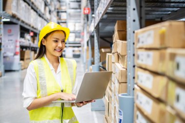 Asyalı mühendis kadın dizüstü bilgisayarda depo, lojistik ve ticari ihracat geçmişine sahip raflardaki lojistik kutuları işaretlemek için hisse senedi bilgilerini kontrol ediyor