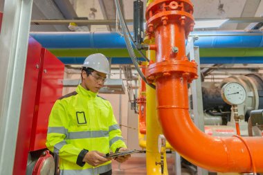 Kazan dairesinde çalışan gözlüklü Asyalı mühendis ısıtma sistemi ekipmanlarının teknik verilerini kontrol ediyor.