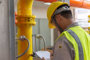 Kazan dairesinde çalışan gözlüklü Asyalı mühendis ısıtma sistemi ekipmanlarının teknik verilerini kontrol ediyor.