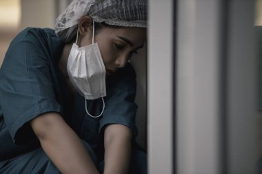 Depresyonda olan Asyalı kadın hemşire hastane zemininde otururken maske takıyor.