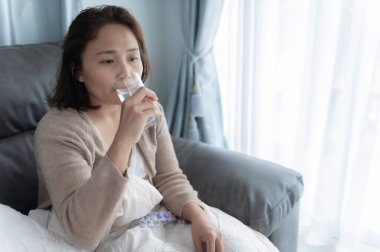 Asyalı hasta kadın kanepede oturur, evde kalır, kadın kendini kötü hisseder, uzanıp dinlenmek ister, ilaç alır.