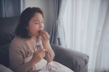 Asyalı hasta kadın kanepede oturur, evde kalır, kadın kendini kötü hisseder, uzanıp dinlenmek ister, ilaç alır.