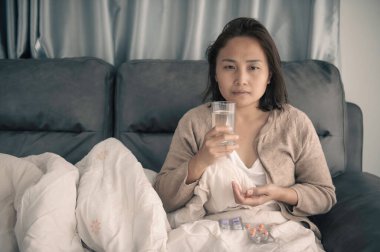 Asyalı hasta kadın kanepede oturur, evde oturur, kadın kendini kötü hisseder, uzanmak ve dinlenmek ister, yüksek ateş, covid-19 omikron gerilimi